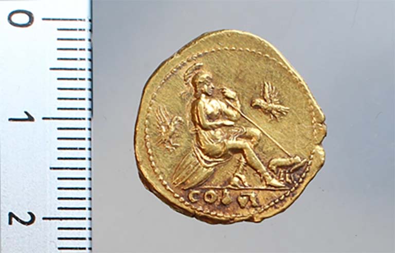 Aureus of Vespasian. One of the gold coins, abandoned in the middle of the skeletons.
77-78 AD Personification of Rome, the left foot, inwards, rests on a helmet. 
At the feet, on the right, the she-wolf breastfeeding Romulus and Remus.
Photo © S. Zanella/S. De Rosa – EFR.
Aureus de Vespasien. Une des monnaies en or, abandonnée au milieu des squelettes.
77-78 ap. J.-C. Personnification de Rome, le pied gauche, vers l’intérieur, repose sur un casque. 
Aux pieds, à droite, la louve allaitant Romulus et Remus.
Photo © S. Zanella/S. De Rosa – EFR.
See Pompei- le secteur de la Porte d’Herculanum à Pompéi Centre Jean Bérard
