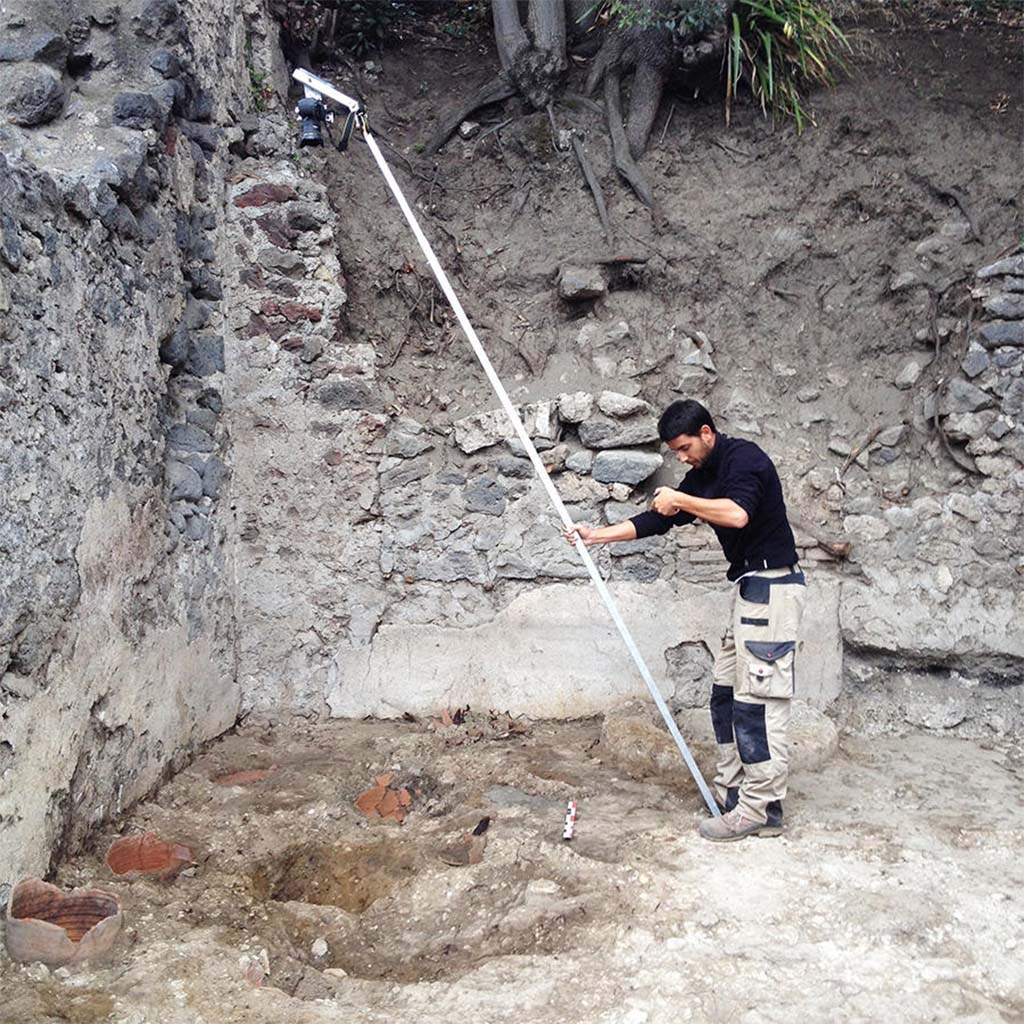HGE28 Pompeii. 2014. Bastien Lemaire photographs the locations of potter’s wheels using a pole. Photo © Guilhem Chapelin.
According to Laetitia Cavassa, after removing the modern rubble, four circular cavities were unearthed, three of whose walls consisted of fragments of cut amphoras; these were the remains of the potter’s wheels. This was a major discovery, as it illustrated through archaeological remains what we already knew, but only with regard to frescoes. Among the rare representations from the Roman period illustrating potter’s wheels at work, two are mural paintings discovered in Pompeii. Now we have these Pompeian wheels. While these traces are fleeting, they indicate the location of the master turners as well as the how these wheels worked. We noted the presence of a central axis made of wood on which the turning wheel was placed. The entire chain of operation has now been illustrated, from the shaping of vases, unfired goblets, and the firing of vases in kilns. 
See Laetitia Cavassa, 2019. Tracing Back the Potters of Pompeii.  Article in English
HGE28 Pompeii. 2014. Bastien Lemaire photographie les emplacements des tours de potiers à l’aide d’une perche. Photo © Guilhem Chapelin.
Rapidement, après avoir enlevé les déblais modernes, nous mettons au jour quatre cavités circulaires dont la paroi est constituée, pour trois d’entre elles, par des fragments d’amphores taillées : il s’agit des vestiges des tours de potiers dont nous cherchions les emplacements. Nous les avons enfin trouvés.
La découverte est de taille : elle vient illustrer par des traces archéologiques ce que nous ne connaissions jusqu’alors que par des fresques. Parmi les rares représentations d’époque romaine illustrant des potiers en train de travailler, deux sont des peintures murales découvertes à Pompéi. Maintenant, nous avons les tours pompéiens. Ces traces, bien que fugaces, indiquent la position des maîtres-tourneurs ainsi que le fonctionnement de ces tours de potiers. On constate la présence d’un axe central en bois sur lequel était placée la roue du tour. Toute la chaîne opératoire est désormais illustrée, de la mise en forme des vases, en passant par les gobelets crus, jusqu’à la cuisson des vases dans les fours.
Voir Laetitia Cavassa, 2019. Sur la trace des potiers de Pompéi.   Article en français