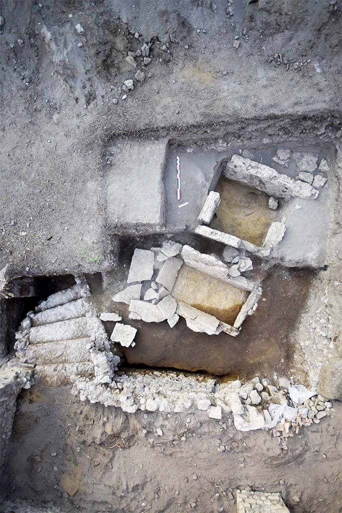 HGE31B with one side missing in 2015. HGE31A is below and the still to be uncovered HGE32A above.
HGE30 is at the bottom with the stairs of the shop at the bottom left.
Photo © B. Lemaire, EFR/CNRS.
Separated by 0.60 m from the previous one and nearly 0.50 m from the third, this grave is completely filled by reworked fill (Figs. 15 and 23). Only three Sarno limestone slabs are preserved and although the north-western one is missing, the space thus delimited is 1.44 m from north-east to south-west and nearly 0.75 m restored from north-west to south-east. The slabs measure between 0.92 and 0.99 m in height (up to 32.30 m s.l.m.) and only the one in the north-east measures only 0.62 m, but is set on wedge stones of about 10 cm. (30102). 
This burial structure was excavated in the modern period, as evidenced by the levels of fill found inside. It is only a single fill. Finally, the space observed was over-excavated by our predecessors, deeper than the installation levels, making it possible to observe the foundation of the slabs and even the wedging elements of one of them. No original floor and fill levels could be observed, nor any elements relating to the remains of the deceased or to offerings.
See CEFR 1581 or para 46 and 47 below