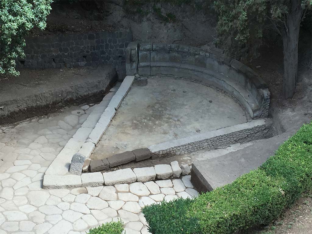 NGH Pompeii. 2015. Schola tomb after clearance showing previously covered tomb and basalt block roads. 
The continuation of ancient Via di Nola into unexcavated area is on the left.
The area immediately outside of Porta Nola (the Nolan Gate) was first cleared in 1907–8. 
Since then, soil had once again accumulated opposite the gate, re-burying the tomb. 
In 2015 the Porta Nola Project has cleared the basalt block roads and brought the monument back to light.
Excavation inside the monument in front of the benches revealed a thick layer of floor preparation for the pavement which has since disappeared.
See BSR Pompeii Project Porta Nola Necropolis 2015 Excavations