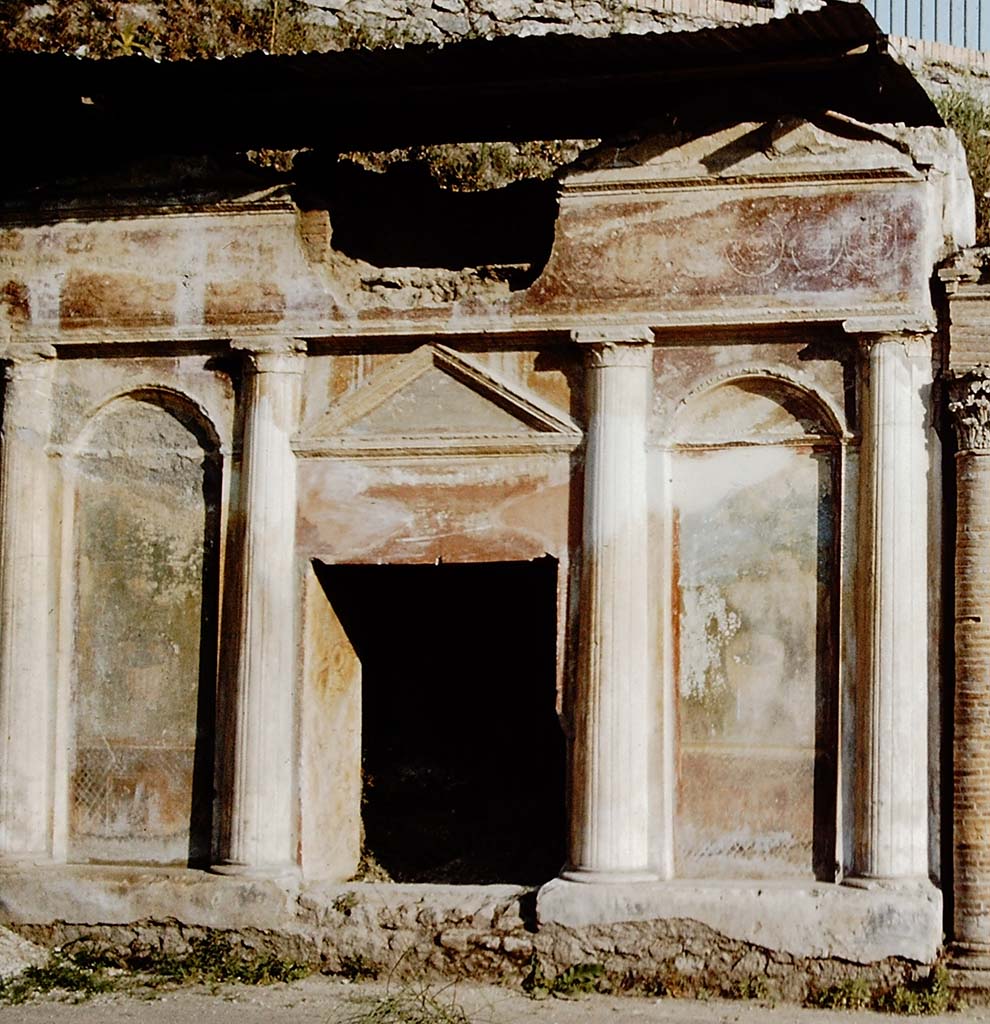 Pompeii Porta Nocera. 1959. Tomb 19ES. Richly decorated front of tomb with four columns, two top pediments, a lunette and stucco bordered panels. 
The front is richly decorated with garden paintings showing fences, marble crater fountains and shrubbery at the top of which are birds. 
Above the columns and below the top pediments are alternating and connected spirals of clovers and palmettes on a red background.
See D’Ambrosio, A. and De Caro, S., 1983. Un Impegno per Pompei: Fotopiano e documentazione della Necropoli di Porta Nocera. Milano: Touring Club Italiano. 19ES.
Detail from photo by Stanley A. Jashemski.
Source: The Wilhelmina and Stanley A. Jashemski archive in the University of Maryland Library, Special Collections (See collection page) and made available under the Creative Commons Attribution-Non-Commercial License v.4. See Licence and use details.
J59f0365