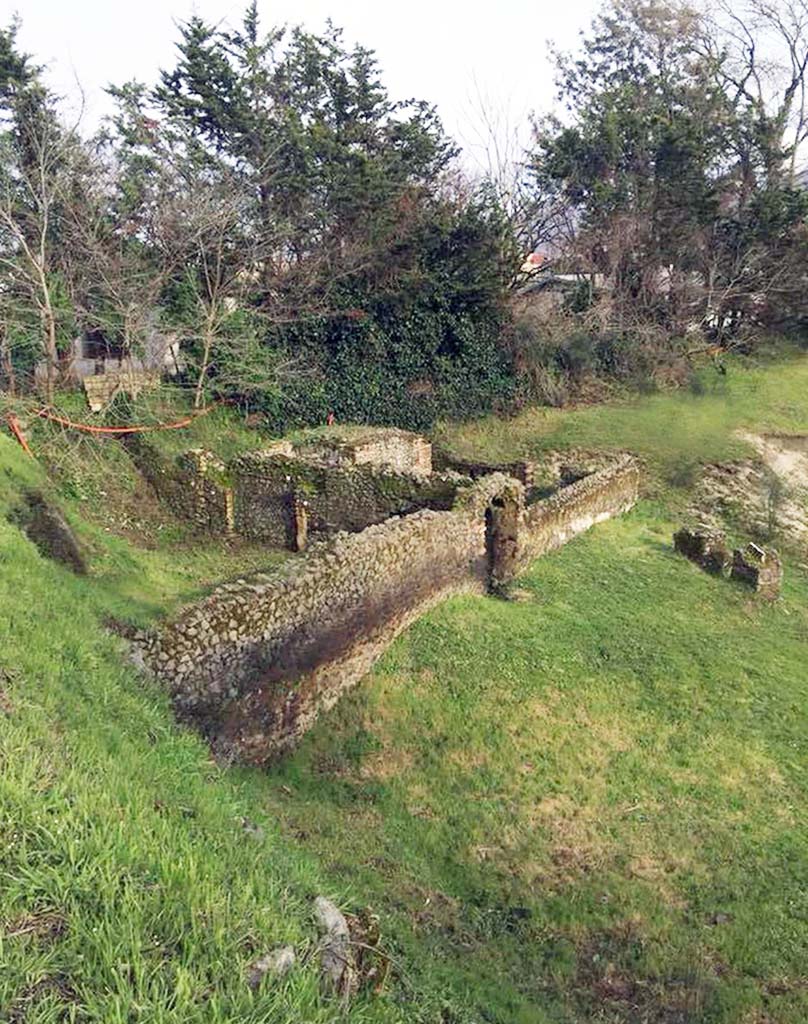 Pompeii Porta Sarno Necropolis. March 2018. Tomb area B, a funerary site with a monument of type “colonnato” is on the left with an arched entrance. 
Area A is the funerary site with a monument on a podium behind tomb B, with its entrance in the far end wall.
The grassed Area J, in front of tomb B, is the space between the funeral precincts and the area of the Samnite necropolis.
The separate stones on the far right are the area H, the Praetorian tombs area.