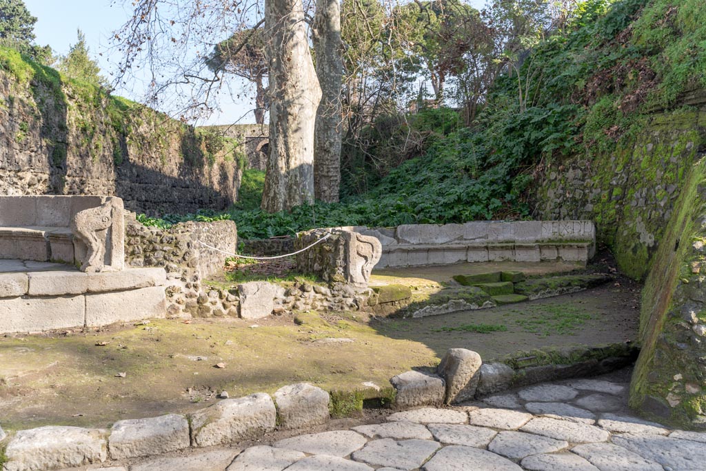 SGD Pompeii, south end on left. January 2023. Looking east towards two schola tombs, with SGF, on right. 
The entrance to the enclosure behind the tomb is in the centre. 
In front of the entrance the road surface, a drain set in the kerb, and the steps in the centre front of the tomb, are visible. 
Photo courtesy of Johannes Eber.