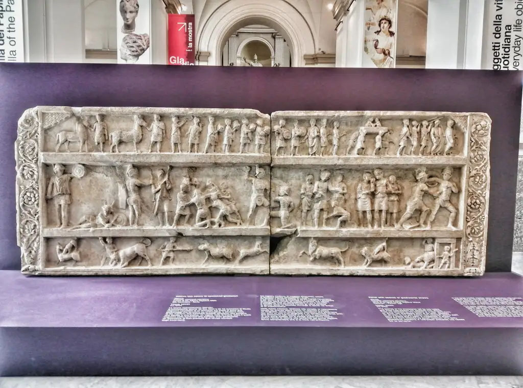 SG6 Pompeii. 2021. Gladiatorial relief with hunting scenes from a tomb at Porta Stabia. The background gives an indication of its large size.
According to Emmerson, this relief measures more than 4 metres long and 1.5 metres high.
See Emmerson A., 2010. Reconstructing the Funerary Landscape at Pompeii's Porta Stabia: Rivista di Studi Pompeiani 21, pp. 78, fig. 1.
According to Osanna, in view of what remains and the visible traces, as well as archive research that is still ongoing, it is more than likely that the top of the tomb SG6, damaged by the construction of the San Paolino building in the nineteenth century, was completed with the gladiator relief preserved in MANN. If you evaluate the role that gladiatorial shows and venations have in praise, it can be assumed that the well-known relief with gladiatorial scenes and hunts with animals kept in Mann was placed there.
The relief has dimensions compatible with the monument SG6, as its length is about 4 metres long, and would respond well in the theme to the role of the deceased as an extraordinary organizer of games. 
Moreover, the relief, discovered by the superintendent Avellino in the 1840s, was found out of place (from the damage evidently suffered by the monument in the construction of San Paolino), precisely in the area of Porta Stabia.
Now in Naples archaeological Museum. Inventory number 6704.
SG6 Pompeii. 2021. Rilievo gladiatore con scene di caccia da una tomba a Porta Stabia. Lo sfondo dà un'indicazione delle sue grandi dimensioni.
Secondo Emmerson, questo rilievo misura oltre 4 metri di lunghezza e 1,5 metri di altezza.
Vedi Emmerson A., 2010. Reconstructing the Funerary Landscape at Pompeii's Porta Stabia: Rivista di Studi Pompeiani 21, pp. 78, fig. 1.
Secondo Osanna, in considerazione di quanto resta e delle tracce visibili, oltre che delle ricerche di archivio tuttora in corso, è più che verosimile che la parte superiore della tomba SG6, danneggiata dalla costruzione dell’edificio di San Paolino nell’Ottocento, fosse completata con un rilievo conservato al MANN. Se si valuta il ruolo che gli spettacoli gladiatori e le venationes hanno nell’elogio, si può ipotizzare che fosse ivi collocato il noto rilievo con scene gladiatorie e di caccie con animali conservato al Mann. 
Il rilievo ha infatti dimensioni compatibili con il monumento SG6, lungo com’è circa 4 m., e risponderebbe bene nel tema al ruolo del defunto di straordinario organizzatore di giochi. 
Inoltre, il rilievo, scoperto dal soprintendente Avellino negli anni ’40 del XIX secolo, fu rinvenuto fuori posto (per i danni subiti dal monumento evidentemente per la costruzione di san Paolino), proprio nell’area di porta Stabia.
Ora al Museo archeologico di Napoli. Numero di inventario 6704.