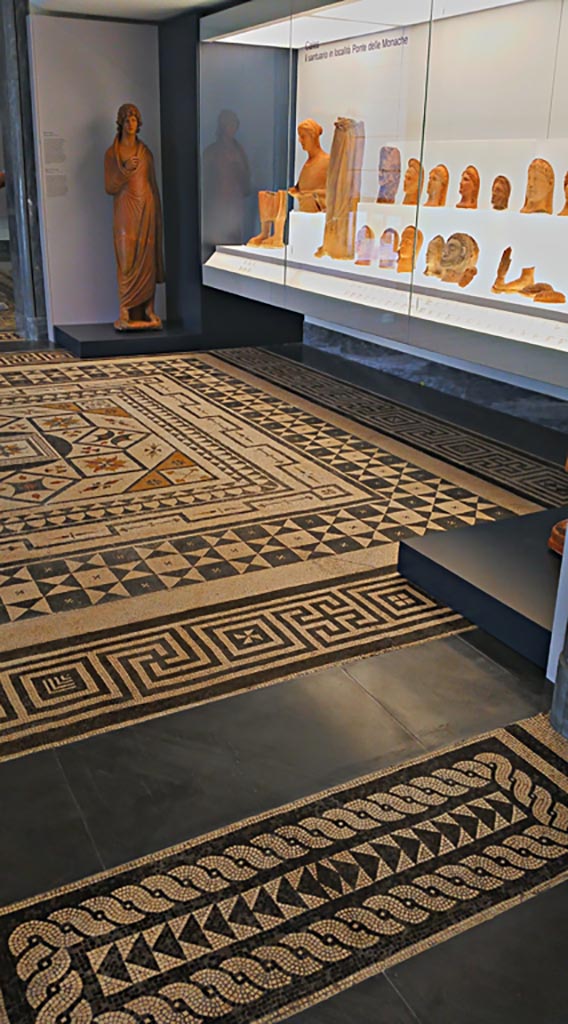 Villa Urbana, Varano, Stabiae. July 2019.
Mosaic floor as seen in room on First Floor in Naples Museum.
Photo courtesy of Giuseppe Ciaramella. 
According to Pisapia –
“Of the four rooms that were paved with mosaic floors, the designs that La Vega made of two of these, lost, nos, 122 and 123, are shown in fig.19, while Ruggiero states that the other two are in Real Museo Borbonico and pave the first and second rooms of the Santangelo Collection.
Of this information, however, only part is correct, since, in the first room of the aforementioned Collection, it has been recognised that there is a mosaic from Lucera, while in the second room it is the floor that we indicate as no.124.”
(Dei quattro ambienti che erano pavimenti a mosaico si riporta alla fig.19 i disegni che il Vega fece di due di essi n.122 e 123 perduti, mentre il Ruggiero afferma che gli altri due sono al Real Museo Borbonico e pavimentano la I e la II sala della Collezione Santangelo.
Di questa notizia, però, soltanto parte era esatta, poiché, nella I sala della Collezione citata e stato riconosciuto esservi un mosaico da Lucera, mentre nella II sala e il pavimento che indichiamo con il no.124).
See Pisapia, M. S. 1989, in Mosaici antichi in Italia, Regione prima. Stabia, Roma, (p.67-70, no.124).
According to Ruggiero –
“Di questo edificio Avanza solamente una copia della pianta (senza il riscontro dei numeri relativi al Giornale perduto) e i musaici dei pavimenti di quattro stanze disegnati dal la Vega; due dei quali si pubblicano con la detta pianta nella Tav. XVIII e due altri si tralasciano, potendosi vederli nel Museo nazionale, l’uno nel mezzo della prima sala dei vasi e l’altro parimente nel mezzo della seconda sala della collezione Santangelo.”
(Of this building remains only a copy of the plan (without the confirmation of the numbers relating to the lost Giornale) and the mosaics from the floors of four rooms drawn by La Vega; two of which are published with the said plan in Tav. XVIII and two others have been left out, as they can be seen in the National Museum, one in the middle of the first room of vases and the other equally in the middle of the second room of the Santangelo collection.)
See Ruggiero, M. (1881). Degli scavi di Stabia dagli 1749 al 1782. (p.356).