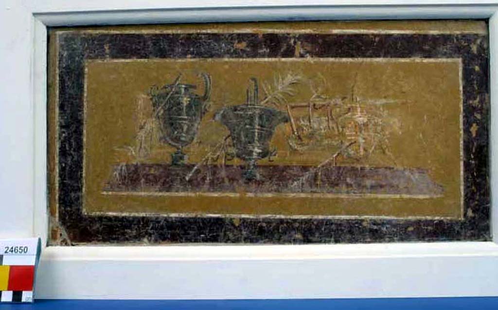 Villa della Pisanella, Boscoreale. Fresco fragment. Decorative compartment with representation of three vases with palm tree branches. 
According to De Cou, the first vase has a wide lip and short neck and is painted to represent a metal surface with raised rings. On the left side of the lower part of the body there is a ring handle with ornamental attachment, a projecting head like that of a horse, and on the stem a festoon of ribbon.
The second vase has flaring sides and a high base and is also painted to represent a metal surface with raised rings. 
The third vase, standing some distance to the right, is shaped like the first but only drawn in outline with raised bands in the centre like the others.
Between the second and third vases is a mat on which is a table with three straight legs visible and an object on top. 
At the right hand end is another table on which an object resembling a cup with a handle is sketched.
See De Cou F. Antiquities from Boscoreale in Field Museum Of Natural History (January 1, 1912), p. 166-7.
Photo © Field Museum of Natural History - CC BY-NC.
Now in the Field Museum, inventory number 24650. See in Field Museum