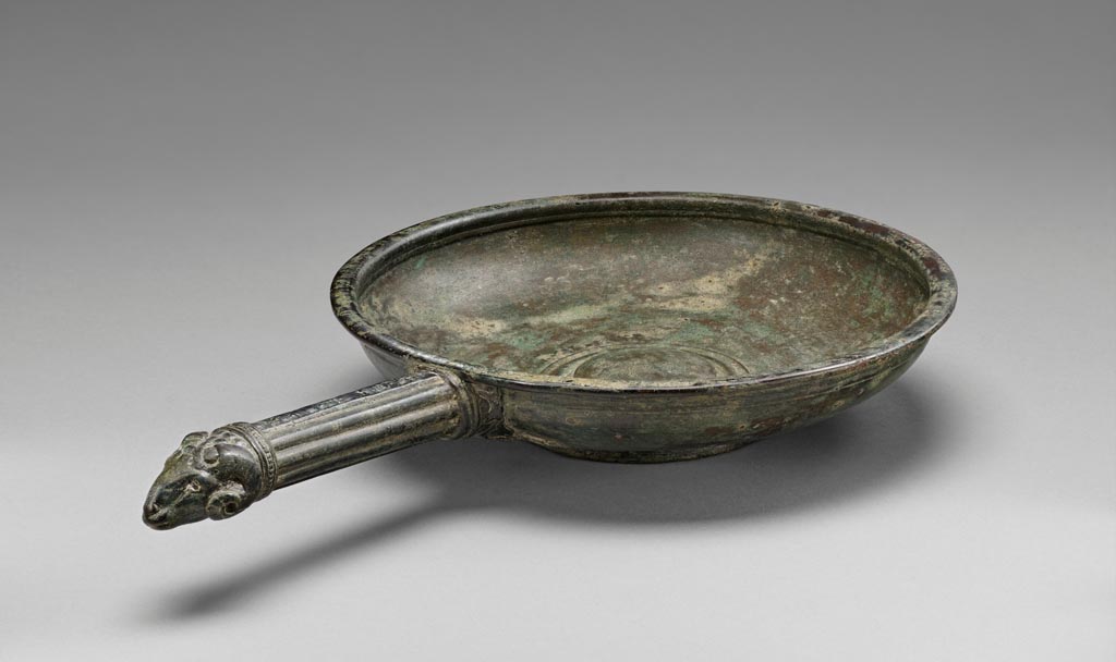 Discovered in a villa in Boscoreale. Bronze patera with ram’s head handle.
Digital image courtesy of the Getty's Open Content Program. Now in the Getty Museum, inventory number 72.AC.138.
The interior and exterior of this bronze patera (shallow bowl) are undecorated, except for a series of concentric bands on the underside of the foot. The tip of the fluted handle is fashioned in the shape of a ram’s head. The attachment that affixed the handle below the rim is now partly missing.
Vessels of this type were used in the home for religious rituals and to hold water for handwashing during banquets. Slaves would pour water from an oinochoe (pitcher) over the hands of participants, catching the cascade in a patera held underneath. This patera was discovered in a villa at Boscoreale or in the vicinity, although the exact findspot is unknown. The owners of the luxurious homes in this area used finely shaped vessels decorated with a variety of images, including mythological figures, theatre masks, animal heads and feet, and floral ornaments.