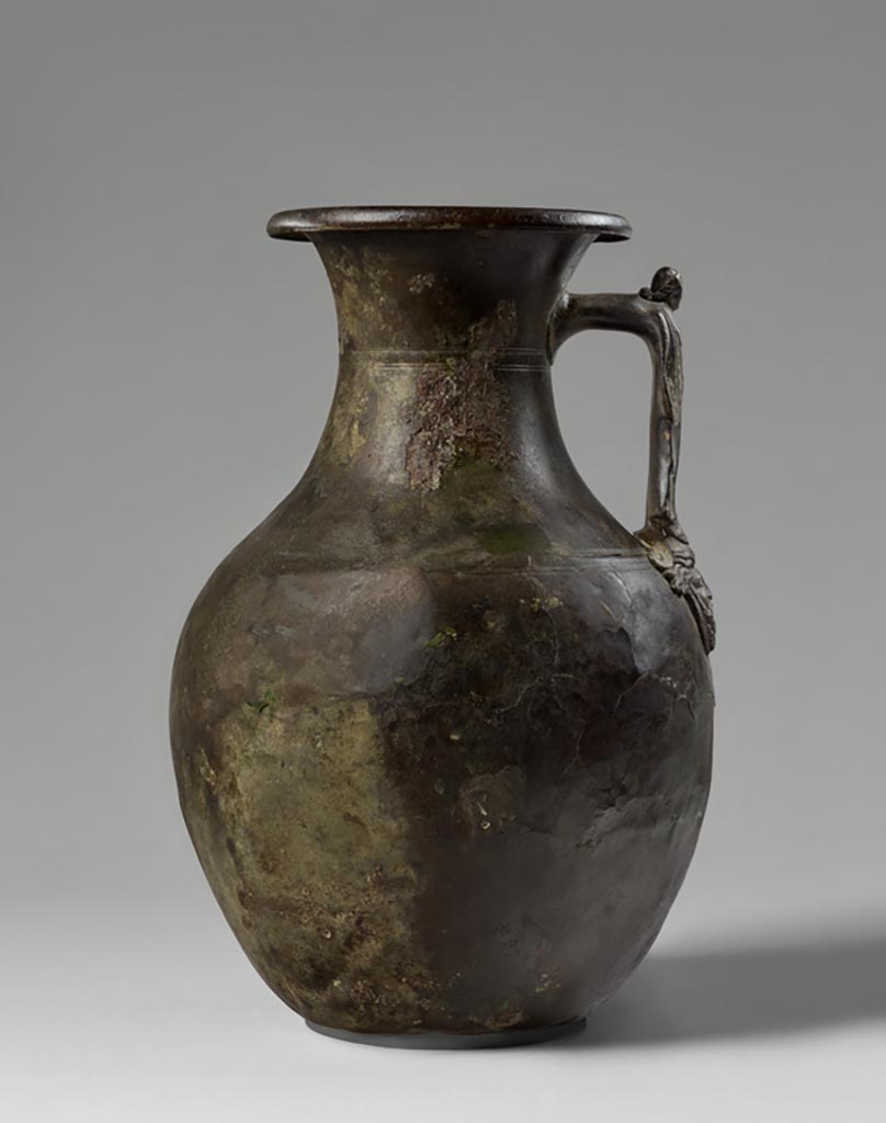 Discovered in a villa in Boscoreale. Bronze amphora with standing female figure and satyr mask.
Digital image courtesy of the Getty's Open Content Program. Now in the Getty Museum, inventory number 72.AC.143.
Aside from two pairs of incised lines on the shoulder and neck of the vessel, the handle, which takes the form of a standing female figure, provides the only decoration on this amphora (storage jar). She wears an ankle-length garment and mantle and holds her arms at her sides. The attachment plate upon which she stands is shaped as a mask depicting a bearded satyr wearing a fillet. 
Satyrs were frequent companions of the wine god Bacchus, and vessels of this type were often used to hold wine at banquets. This amphora was discovered in a villa at Boscoreale or in the vicinity, although the exact findspot is unknown. The owners of the luxurious homes in this area used finely shaped vessels decorated with a variety of images, including mythological figures, theatre masks, animal heads and feet, and floral ornaments.