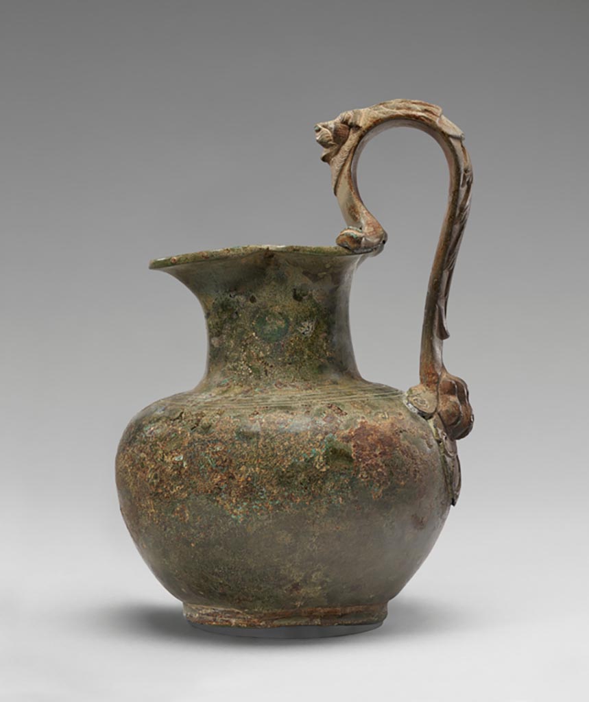 Discovered in a villa in Boscoreale. Bronze oinochoe (pitcher) with lion handle.
Digital image courtesy of the Getty's Open Content Program. Now in the Getty Museum, inventory number 72.AC.146.
The shoulder of this otherwise plain oinochoe (pitcher) is decorated with two pairs of incised bands. Parts of the high, arched handle, however, take the form of a lion. A lion’s head in relief appears above the vessel’s mouth. The rotellae, small protrusions on either side of the handle where it joins the rim, are fashioned as the stylized forelegs of the feline. At the base of the handle is a lion’s paw. Floral motifs run along the outer face of the handle.
Vessels of this type were used in the home for religious rituals and to hold water for handwashing during banquets. Slaves would pour water from an oinochoe over the hands of participants, catching the cascade in a patera (shallow bowl) held underneath. This oinochoe was discovered in a villa at Boscoreale or in the vicinity, although the exact findspot is unknown.