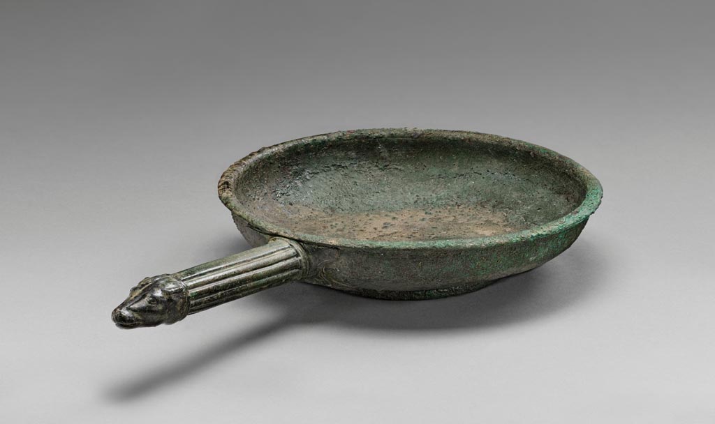 Discovered in a villa in Boscoreale. Bronze patera with dog’s head handle.
Digital image courtesy of the Getty's Open Content Program. Now in the Getty Museum, inventory number 72.AC.134.
The interior and exterior of this bronze patera (shallow bowl) are undecorated. A series of concentric bands decorate the underside of the foot. The tip of the fluted handle is fashioned in the shape of a dog’s head. At the join to the bowl is a relief attachment comprised of floral ornaments.
Vessels of this type were used in the home for religious rituals and to hold water for handwashing during banquets. Slaves would pour water from an oinochoe (pitcher) over the hands of participants, catching the cascade in a patera held underneath. This patera was discovered in a villa at Boscoreale or in the vicinity, although the exact findspot is unknown.