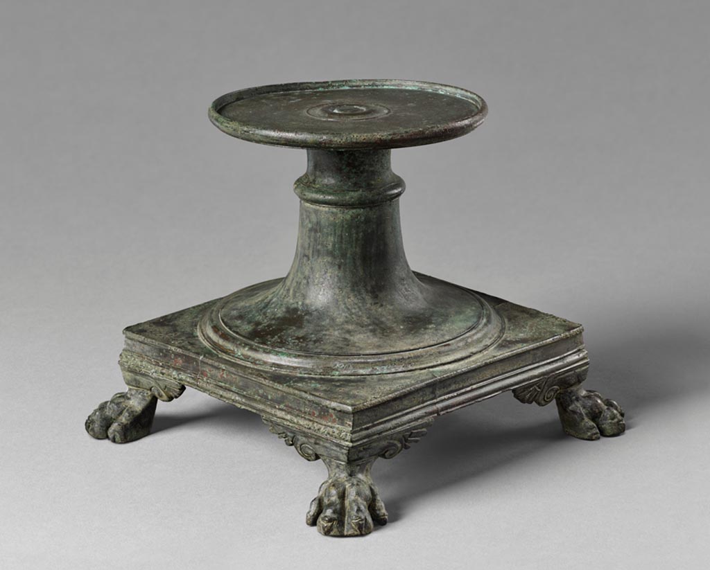 Villa in Boscoreale. Bronze stand.
Perched on four small lion’s paws edged with palmettes, this stand comprises a square plate; a tapering cylindrical riser with a flaring base and a raised collar; and a flat, broad, round plate on which a separate vessel—perhaps a large krater, a bowl used for mixing wine and water at drinking parties - would stand.
Like the Greeks before them, the Romans diluted their wine with water. This practice gave rise to a variety of containers and utensils designed for the mixing and serving of wine. As part of an ornate table service, a krater elevated on a stand like this one would have made a striking presentation.
As was frequently the case with Roman bronze implements, the various parts of the stand were cast separately and then assembled.
Digital image courtesy of the Getty's Open Content Program. Now in the Getty Museum, inventory number 72.AC.135.
