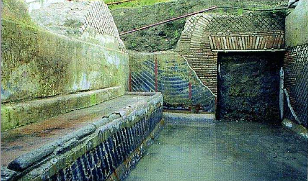 Complesso dei triclini in località Moregine a Pompei. Excavation photo. The kitchen with the large bench and the doorway leading to the triclinium area. 
The kitchen (F) is large, 4.40 x 7.05 meters wide with the long and wide cooking bench against the eastern wall (4.75 x 1.40 metres); a large door to the south allowed one to reach the triclinium area, while another door to the north, on the west wall, allowed one to reach the uncovered service area (fig. 5). The cocciopesto floor has a slope towards the north-east corner of the room, here, just at the corner, a sort of pillar contains a large gutter made of terracotta tubules (about 22 cm in diameter) which probably collected part of the roof water. The decoration of the walls with a black background is very simple as is characteristic of rustic environments as well. the lararium scene in the centre of the west wall: all the walls are divided into panels delimited by red bands, alternately decorated with simple white streaks now arched now oblique. Likewise, the walls of the cooking counter. At the centre of the west wall is a large lararium painted with scenes of sacrifice on a white background. In the foreground, in the centre, we see an altar to which two long snakes approaching, rich in details as regards the scales. In the background, also in the centre, there is a priest, head veiled, dressed in a white mantle with red borders. On the sides two attendants with ivy crowned heads and short white robes act as helpers.
See Nappo, S. C., 2005. I triclini di Murecine uso ed interpretazione, p. 59, fig. 5.