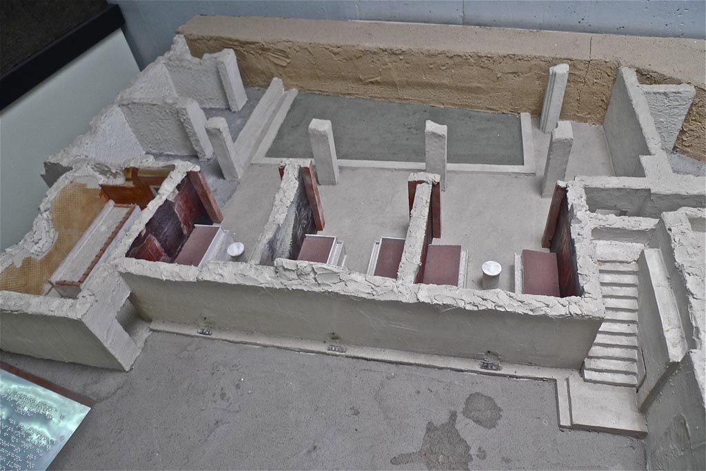 Complesso dei triclini in località Moregine a Pompei. July 2010. Model looking south. The kitchen with a 3.75m long bench is on the left in this photo.
Photo courtesy of Michael Binns.
The main door from the kitchen to the triclinia is at the front and a secondary door appears to lead to the rear where the model is incomplete.
Although complete with everything, at the time of the eruption the kitchen was not in operation and was used as a deposit. In fact, around 200 marble slabs (1.80 m × 0.75 m) were found neatly stacked in it, presumably to be used to decorate the unfinished bath complex.
According to Perna, Antonelli and Lazzarini, in an archaeometric analysis of the greco scritto marble slabs from the Edificio dei Triclinii, discovered at Murecine in Pompeii’s suburbs in 1999–2000, a total of 14 samples were collected and subjected to standard X-ray diffraction (XRD), optical microscopy (OM) on thin sections and stable isotope ratio analysis (SIRA) to assess the stone’s identity and provenance. The results of this study substantiate the hypothesis (already put forward and based on a macroscopic identification by Perna and Scognamiglio) that the stone originates from the Hasançavuşlar quarries near Ephesus in Asia Minor.
See Simona Perna, Fabrizio Antonelli, Lorenzo Lazzarini. Archaeometric analysis of the ‘greco scritto’ marble slabs from the Edificio dei Triclinii at Murecine. Wiley Online Library.