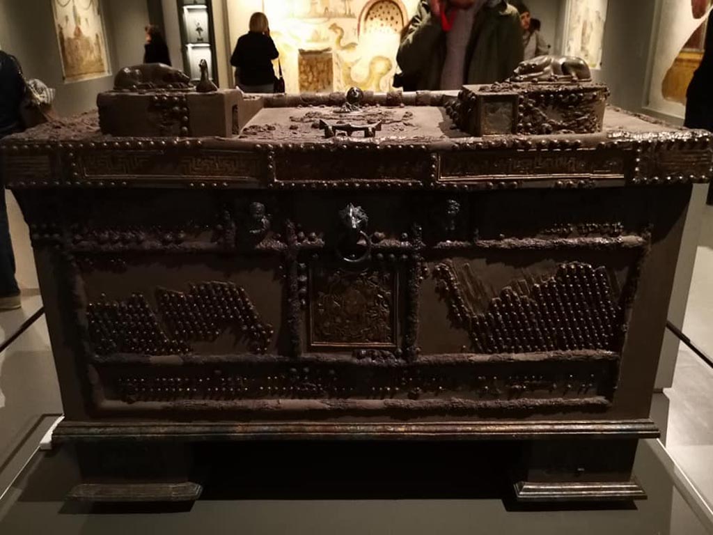 Oplontis, Villa of Lucius Crassius Tertius. December 2019. Strongbox, found in the portico. 
On display in exhibition “Pompei e Santorini” in Rome, 2019. Photo courtesy of Giuseppe Ciaramella.
The inscription crediting three Greek artisans with its creation dates from between the First and Third Centuries BCE, so it was already at least 200 years old when Vesuvius erupted. 
The artisans embellished the strongbox with an elaborate inlaid silver and copper head of the satyr Silenus surrounded by twisting vines, as well as with several sculptural elements – bronze dogs’ heads and a duck, among others – that would together have contributed to a complex locking system, requiring specialized knowledge to undo.