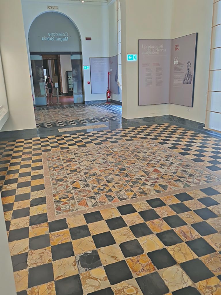 Mosaic in Naples Archaeological Museum, room CXLII (142), looking towards room CXLIII (143). 
June 2024. Photo courtesy of Giuseppe Ciaramella.
According to Miele –
“Around the central panel (from room 13 of the so-called Secondo Complesso) is another floor with alternating slate and antique yellow squares, detached from an apsidal building next to the Second Complex, while the large squares from the same floor are placed in room CXXXVIII.”
See Miele, F: Un Complesso di pavimenti a mosaico e sectilia nel Museo Archeologico Nazionale di Napoli, (p.65-69).
In: Museo Archeologico Nazionale di Napoli, “La Collezione Magna Grecia”.