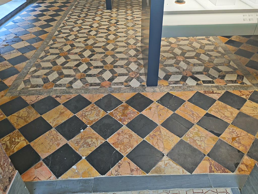 Mosaic in Naples Archaeological Museum, room CXXXVIII (138). June 2024. Photo courtesy of Giuseppe Ciaramella.
According to Miele –
“In the centre of the floor is a floor detached from the Villa Imperiale on Capri, surrounding it are the larger alternating slate and antique yellow squares, detached from an apsidal building next to the Second Complex.”
According to La Vega –
28 Ottobre 1775.
In Stabia si e quasi del tutto scavata la stanza notata in piu antecedenti rapporti; e questa rettangola e termina da un lato con semicircolo che l’abbraccia da due lati; la parte rettangola e di palmi 29 ed once 5 per palmi 31 ed once 5, ed il semicircolo ha di diametro palmi 48 ¼; le mura che la chiudono erano bene dipinte con fondo rosso, ma ora ne restano in piedi per solo otto o nove palmi d’altezza; il pavimento e di marmo con alternativi quadrati, uno di giallo antico ed altro di lavagnone, ed attorno al margine vi sono delle fasce di pietre di vari colori.
Li quadrati che fanno il pavimento della parte rettangola della stanza sono di lato once 14 e quelli che fanno il pavimento della parte semicircolare sono once 10. Subito che questo pavimento sara intieramente scoperto avvisero Canart che lo mandi a levare…………………
(In Stabia, the room noted in several previous reports has been almost completely excavated: it is rectangular and ends on one side with a semicircle that embraces it on two sides; the rectangular part is 29 palms and 5 onces by 31 palms and 5 onces, and the semicircle has a diameter of 48 ¼ palmi; the walls that enclose it were well painted with a red background, but now they remain standing only 8 or 9 palmis high; the floor is made of marble with alternative squares, one of Antique Yellow and the other of Lavagnone, and around the margin there are bands of stones of various colours.
The squares that make the floor of the rectangular part of the room are 12 inches, and those that make the floor of the semicircular part are 8 inches. As soon as the floor will be completely uncovered, I will advise Canart to lift it…….)
See Ruggiero M., 1881. Degli scavi di Stabia dal 1749 al 1782, Naples, (p.232).