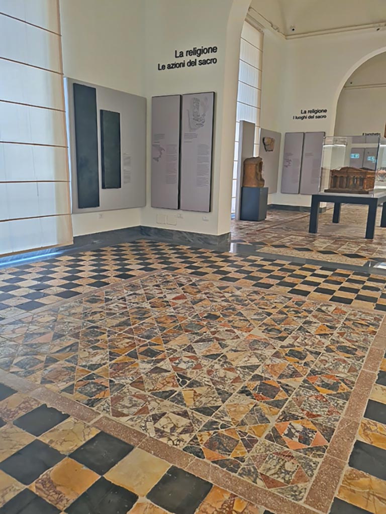 Mosaic floor in second room on first floor of Naples Archaeological Museum. Looking towards third room. 
June 2024. Photo courtesy of Giuseppe Ciaramella. 
According to Naples Museum notice-card –
“With the recent intervention of restructuring of the room ready for the opening of the Collection Magna Grecia, it was decided not to cover the mosaics again and to effect the necessary restoration and maintenance of them to enable the public to see and gain access to them.”
According to Pisapia –
 “Il pavimento è stato riconosciuto in una delle sale del I piano del Museo Nazionale di Napoli, ma è stato strappato assieme a quelli di tutta l’ala, per cui mi è stato impossibile reperire una buona fotografia.” (Note 4 – page 55).
(The floor has been recognized in one of the rooms on the first floor of the National Museum of Naples, but it was removed along with those of the entire wing, so that it was impossible for me to find a good photograph.)
See Pisapia, M. S. 1989, in Mosaici antichi in Italia, Regione prima. Stabia, Roma, Room 104: p.54-55. (Her room 104 corresponds with our Room 13).