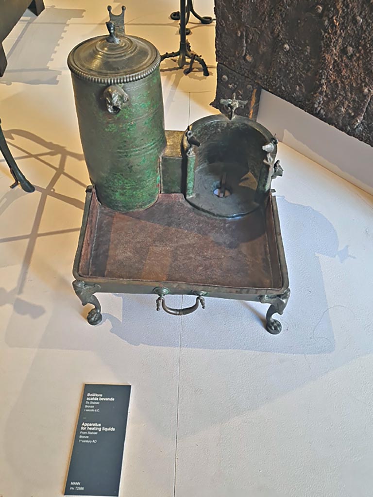 Stabiae, Villa Arianna, October 2023. Room 10, heating appliance, inv. 72986. Photo courtesy of Giuseppe Ciaramella.
On display in “L’altra MANN” exhibition, October 2023, at Naples Archaeological Museum.
According to Pagano and Prisciandaro, this was found in room 10, 23rd October 1758.
Their other references were Rami inediti, fig.198 a and b, Ruesch,1695, Tav. IV, Masseria Irace, 10.
See Pagano, M. and Prisciandaro, R., 2006. Studio sulle provenienze degli oggetti rinvenuti negli scavi borbonici del regno di Napoli. Naples: Nicola Longobardi, 
(p.242).
Sulla destra del fornello un rubinetto, decorato a testa di leone, permetteva al liquido riscaldato di uscire secondo le esigenze. L’ansa del coperchio è a forma di bustino di Mercurio e sulla parete del contenitore è fissata una applique a maschera comica; tre cigni decorano il fornello e tre maniglie “a goccia” sono applicate ai lati del vassoio.
(trans: On the right of the stove a tap, decorated with a lion’s head, allowed the heated liquid to flow out as needed. The handle of the lid is in the shape of a head of Mercury and fixed on the wall of the container is a comic mask. Three swans decorate the stove and three “teardrop” handles are applied to the sides of the tray.)
See Le Collezioni del Museo Nazionale di Napoli, 1986, Rome, De Luca Editore, (p.182-3, no.74), and with reference to MB 1829, V, Tav. XLIV.