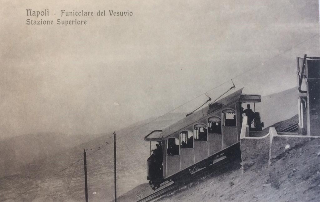 Postcard for Vesuvius Funicular, showing Upper Station. Not dated but must be between 1910 and 1944.
There is a handwritten note on rear – “The 1910 Funicular lasted until 1944”.  Photo courtesy of Rick Bauer.
After the 1906 eruption the funicular was out of action and reconstruction was only completed in 1909.
It reopened in 1910 with two new 5-bay end loading cars, numbered 1 and 2, which seated 16 with 8 standing. 
The car in this photo is car number 1.
The line now had overhead wire and the cars bow collectors to provide electric light, bell signals and a telephone. 
The funicular could now run an after dark service.
The funicular was permanently closed after being destroyed by the Vesuvius eruption of 1944.