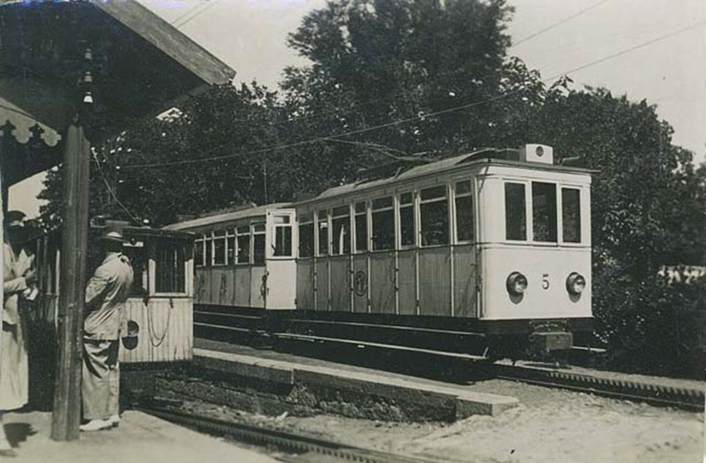 Vesuvius to Pugliano railway. 1934. Hotel Eremo station. Photo courtesy of Rick Bauer.
The same carriages were used for a journey along the whole of the track.
From Pugliano to the power station and from Eremo to the Funicular the trains used their own power and traction was by simple adhesion to the rails.
The rack system of the track is visible in front of the rack locomotives and carriages. 
The rack locomotive on the left was used to push the carriages up the steepest part of the track with up to 25% gradient from the power station to Eremo. 
Traffic had developed to the point where larger cars were needed.
Car number 5 on the right was introduced in the 1920’s and seated 34 passengers who were fully enclosed.
Two other newer cars, numbered 6 and 7 each seated 44 passengers.
The original cars 1 to 3 had seated only 24 people with an additional 6 standing on the platform. They were open above the waistline with curtains for use against wind and rain. A fourth enclosed car had been added when the railway was extended to the relocated and rebuilt funicular base station on 6th January 1913. 
See Smith P, 1998. Thomas Cook and Son’s Vesuvius Railway in Japan Railway and Transport Review March 1998.